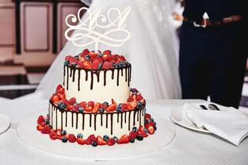Elegant white wedding cake decorated with fresh berries and chocolate glaze with bride and groom on background, copy space