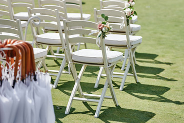 White wooden chairs with rose flowers on each side of archway outdoors, copy space. Empty chairs for guests prepared for wedding ceremony on golf course