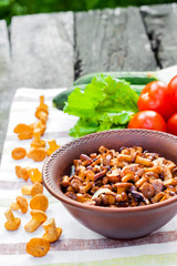 Fried chanterelles with onion in rustic bowl and plate with fresh vegetables for salad on background