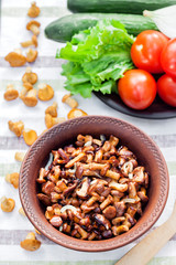 Fried chanterelles with onion in rustic bowl and plate with fresh vegetables for salad on background