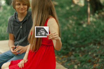 A happy married couple is holding in their hands a snapshot of an ultrasonic study of the baby. Husband hugs a beautiful adult pregnant wife. Second trimester.