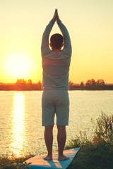 Vertical photo. A young man is standing on the lake at sunset, doing yoga. Ashtanga, ekam, nava. Balance, harmony, balance, concentration, relaxation. asana
