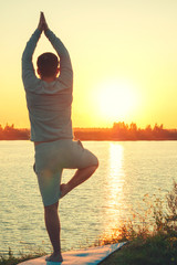 Vertical photo. A young man is standing on the lake at sunset, doing yoga. Stands in a pose of tree a Sathi Yoga. Balance, harmony, balance, concentration, relaxation.