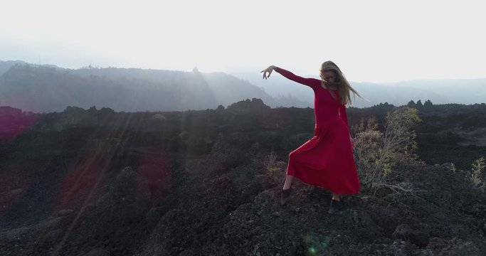 Aerial view blonde young woman in red dress dancing among the black rocky landscape at sunset. 4K