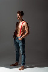 shirtless and handsome man touching red suspenders while standing on grey