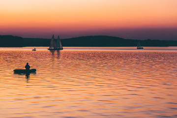 man fishing on a lake from the boat at sunset. silhouette of fishermen. yellow and orange sunset background. sailing boats in water at evening.