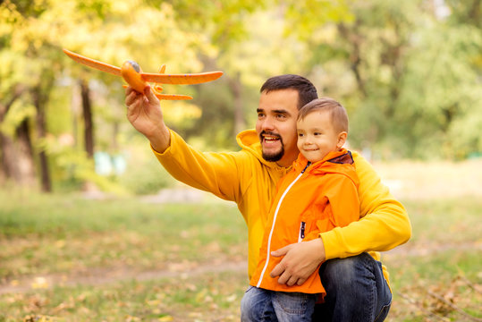 Father and little son play together in autumn park or forest. Dad is embracing child and launching orange toy airplane. Both man and boy are happy and smiling. Family spending good time outdoors