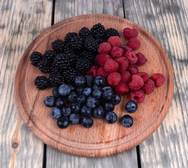 on a wooden table of gray color lies a board on which ripe berries of dark violet blackberry, blueberry and red raspberry are laid