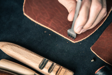 a young tanner on a desktop in a workshop punches holes for stitching on a piece of brown leather
