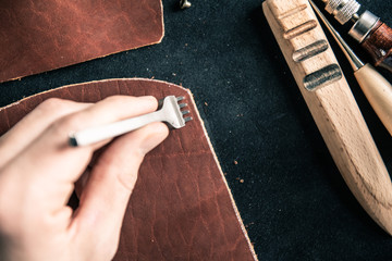 a young tanner on a desktop in a workshop punches holes for stitching on a piece of brown leather
