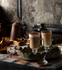 two tall glasses with layered coffee drink with foam and cocoa on top stands on vintage tray on rustic wooden table with copper pot, coffee beans opposite concrete wall, selective focus