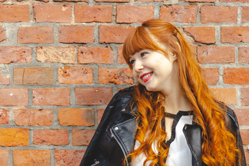 Cute young woman in a black leather jacket with curly red hair and bangs smiles against a brick wall with copy space