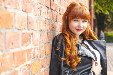 Cute young woman in a black leather jacket with curly red hair and bangs smiles against a brick wall