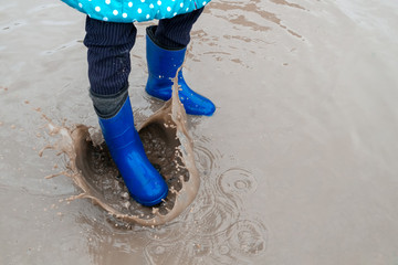 the child is dressed in rubber boots strongly stomps on a puddle. drops of dirty water spray everywhere.