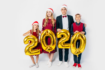 Young happy family with kids wearing Santa hats holding balloons shaped like numbers 2020 on white...