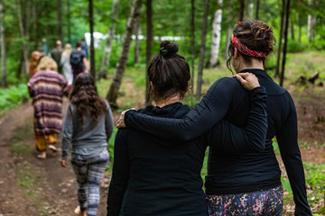 Diverse people enjoy spiritual gathering Two women support each other arm in arm, as they hike a forest trail with a group of individuals experiencing native cultures and traditions.
