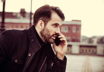 Serious thinking business man in fashion clothing texting sms looking on mobile phone in the hand outdoors autumn background.