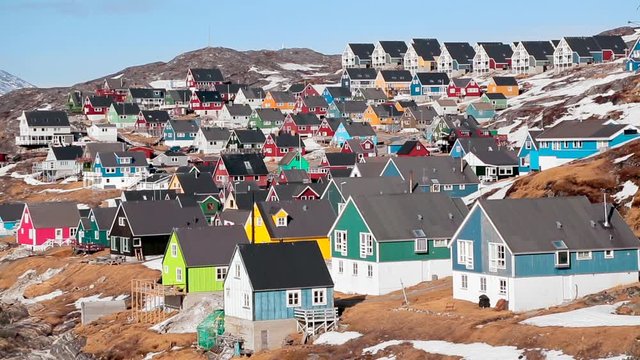 Rows of brightly painted wooden houses climb a bare rocky slope in Nuuk in Greenland with the old spring snow still lying between them. A vivid and cheerful scene of a small Arctic town.