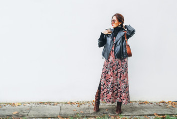 Smiling female dressed boho fashion style colorful long dress with black leather biker jacket with brown leather flap bag posing on the white wall background.