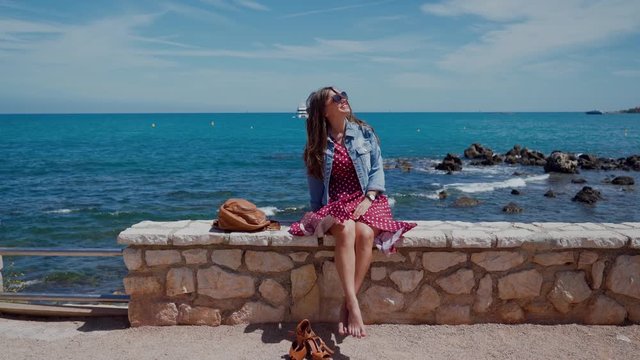 Beautiful young woman with long hair in red summer dress sitting on the stone fence wall, dangling his bare feet. Her shoes stand nearby. Incredible views of the Mediterranean Sea in the background. D