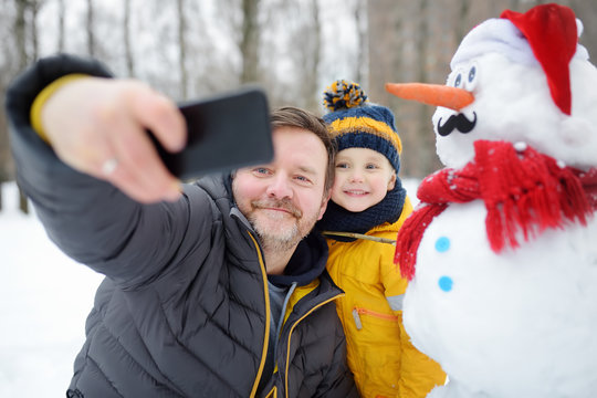 Little boy and his father taking selfie on background of snowman in snowy park. Active outdoors leisure with children in winter.