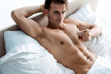 young sexy man lying on bed and holding glass with whiskey