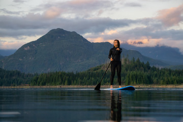 Adventurous Girl on a Paddle Board is paddling in a calm lake with mountains in the background during a colorful summer sunset. Taken in Stave Lake near Vancouver, BC, Canada.