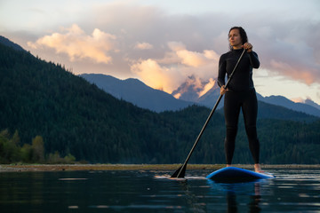 Adventurous Girl on a Paddle Board is paddling in a calm lake with mountains in the background during a colorful summer sunset. Taken in Stave Lake near Vancouver, BC, Canada.