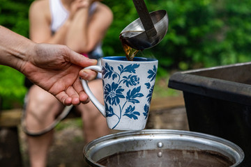 Diverse people enjoy spiritual gathering A close up view on the hands of a person serving healthful and natural tea from a pan over a campfire at a sacred forest clearing.