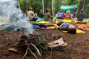 Diverse people enjoy spiritual gathering A close up view on a smoldering camp fire as blurry people are seen in deep meditation and prayer in the background during a mindful forest retreat.
