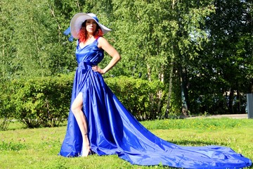 Fototapeta premium a lady in a blue dress and a gorgeous snow-white hat on a green lawn in the Park. a woman in a hat and a long dress with a train on the skirt on the background of green grass and trees in the city