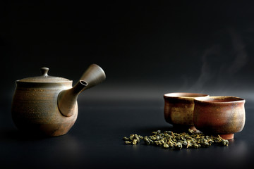 Raw tea leaves, teapots and two tea cups have smoke from the heat on a black background.