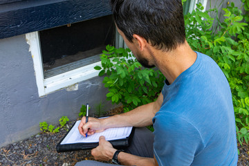 Indoor damp & air quality (IAQ) testing. A close up and high angle view of a building inspector checking for defects near a basement window of a domestic home during an environmental quality exam. © Valmedia