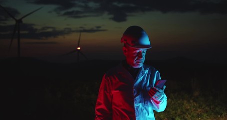 Caucasian engineer in white suit working at wind turbine farm checking smartphone content staying in the field at night. Beautiful scenery in background.