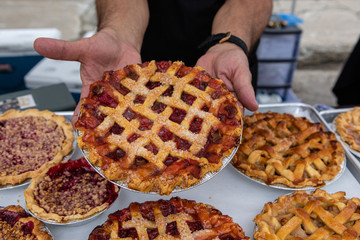 Baked goods at outdoor agriculture fair. A market trader is seen close up, holding a freshly baked fruit tart with pastry topping by a display of freshly baked goods during a farmer's market.