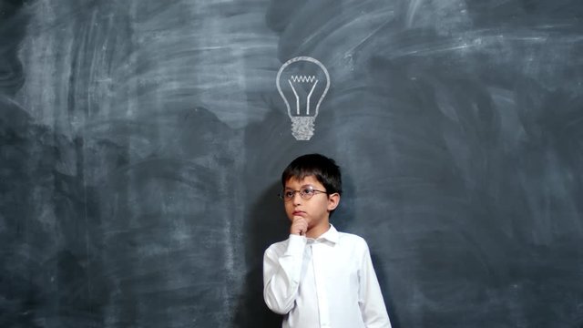 Smart little schoolboy in white shirt and eyeglasses standing against blackboard with animated chalk drawings of light bulb, rubbing his chin and thinking hard about something