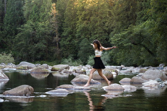 Young Attractive Brunette Woman Hopping Over Rocks Beside A Pristine River In An Evergreen Forest.