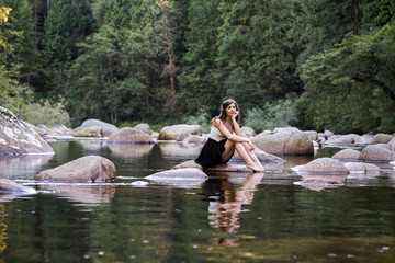 Young attractive brunette woman on a rock next to a pristine river in an evergreen forest.