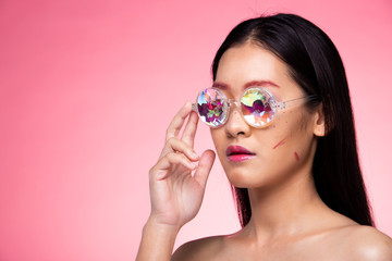 Fashion Model looks at camera for shooting new collection Sunglasses.  Beautiful Asian Woman summer trend make up wear Kaleidoscope Glasses with colorful, studio lighting pink background