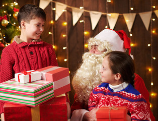 Santa Claus and child boy and girl posing together indoor near decorated xmas tree with lights, they talking, smiling and accepting gifts - Merry Christmas and Happy Holidays!