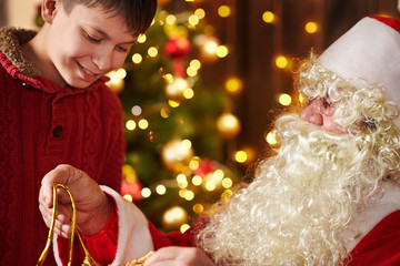 Santa Claus opens a bag and gives gifts to children, sitting indoor near decorated xmas tree with lights - Merry Christmas and Happy Holidays!