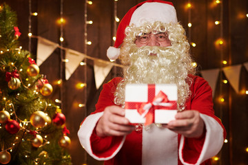 Closeup of Santa Claus hands with gift box, sitting indoor near decorated xmas tree with lights - Merry Christmas and Happy Holidays!