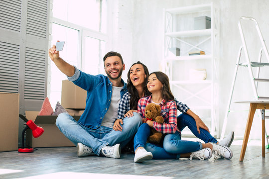 Like in a fairy tale. Young family of father, mother and daughter are sitting on the floor in the a new apartment laughing and taking a selfie together.