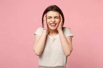 Laughing funny young woman in casual light clothes posing isolated on pastel pink background studio portrait. People lifestyle concept. Mock up copy space. Keeping eyes closed putting hands on cheeks.