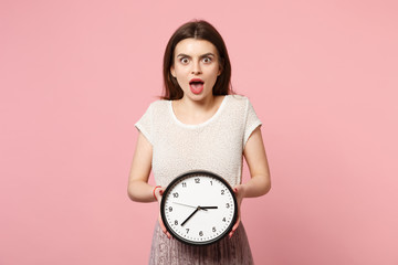 Shocked young woman in casual light clothes posing isolated on pastel pink wall background, studio portrait. People lifestyle concept. Mock up copy space. Holding in hands clock, keeping mouth open.