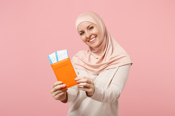 Smiling young arabian muslim woman in hijab light clothes posing isolated on pink background. People religious Islam lifestyle concept. Mock up copy space. Holding passport, boarding pass ticket.