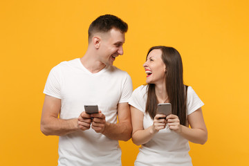 Laughing young couple friends guy girl in white t-shirts posing isolated on yellow orange background. People lifestyle concept. Mock up copy space. Holding using mobile phone, looking at each other.