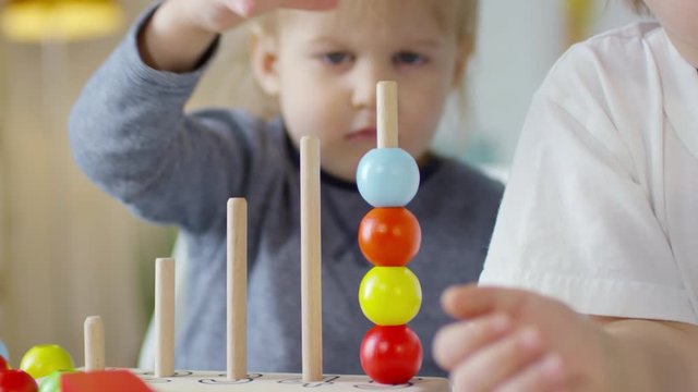 Close up view of cute toddler boy putting colorful beads on wooden stick while playing with educational toys in kindergarten