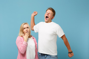 Fototapeta premium Young tired couple two friends guy girl in white pink empty design t-shirts posing isolated on pastel blue background. People lifestyle concept. Mock up copy space. Yawning, covering mouth with hand.