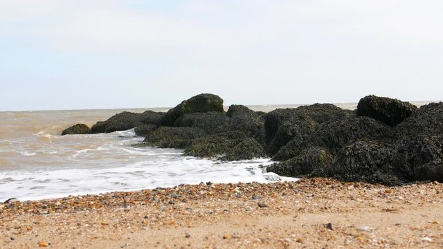 4K Waves against a Rock pool in a British Beach by the sea, sandy rocky Beach with rocks moss and grass. Shoreline with shells, pebbles and sand.